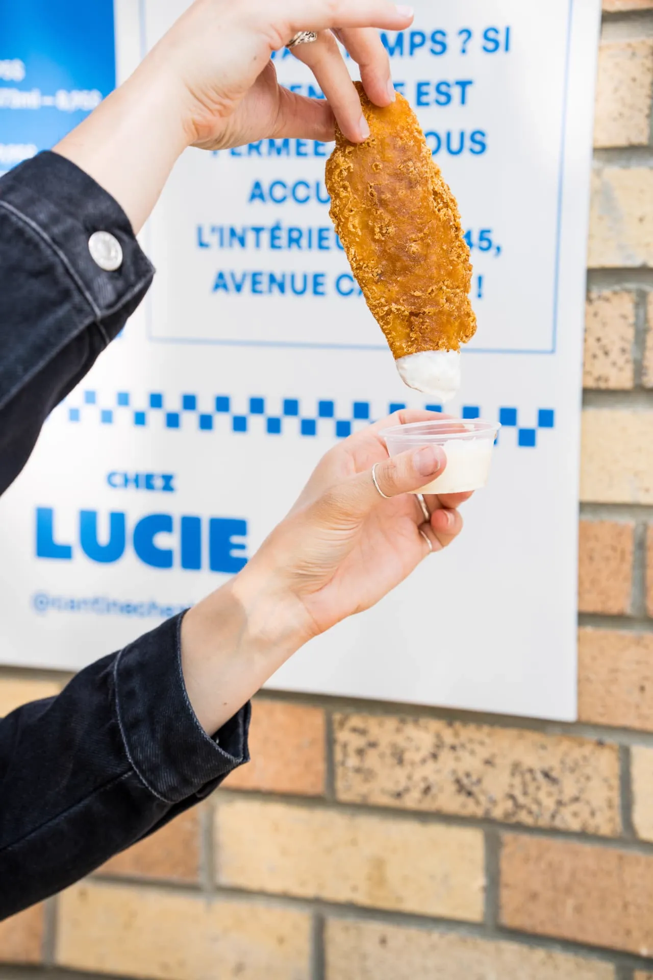 Doigt de poulet trempé dans une sauce devant l'affiche Chez Lucie