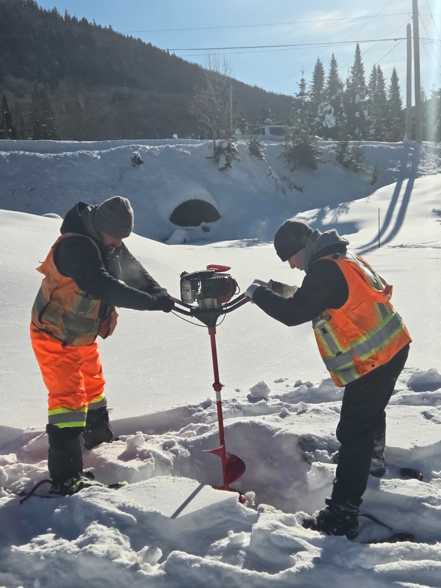 Deux arpenteurs percent la neige avec une tarière motorisée lors d’un relevé hivernal.